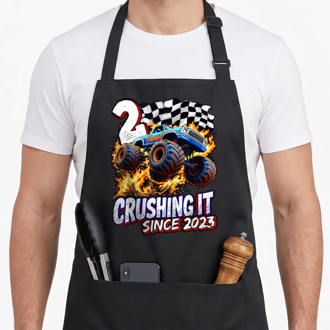 Birthday monster 2nd truck apron - Blue monster truck leaping through fire with checkered flag and CRUSHING IT SINCE — shown on a black cotton canvas apron against a white background