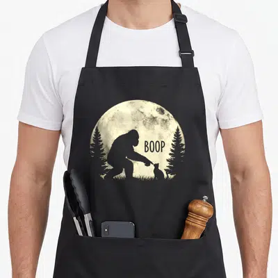Boop cat apron - Bigfoot silhouette touching small cat with BOOP text under textured moon — shown on a black cotton canvas apron against a white background