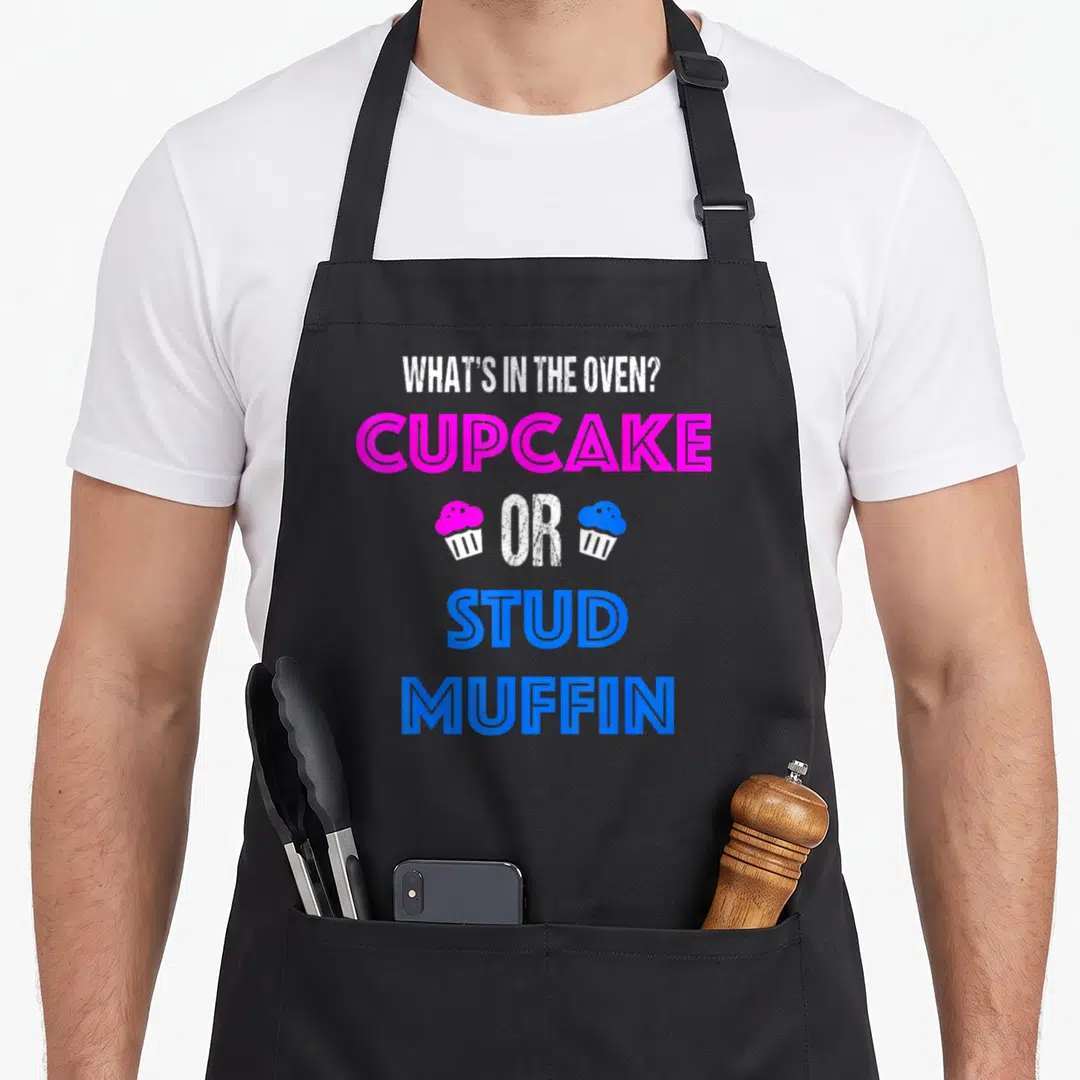 Cupcake muffin man apron - Stacked text reading WHAT'S IN THE OVEN CUPCAKE OR STUD MUFFIN with pink and blue icons — shown on a black cotton canvas apron against a white background