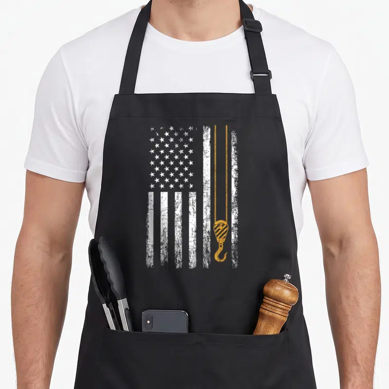 Crane american flag apron - Distressed white USA flag with orange crane hook and cable overlay — shown on a black cotton canvas apron against a white background