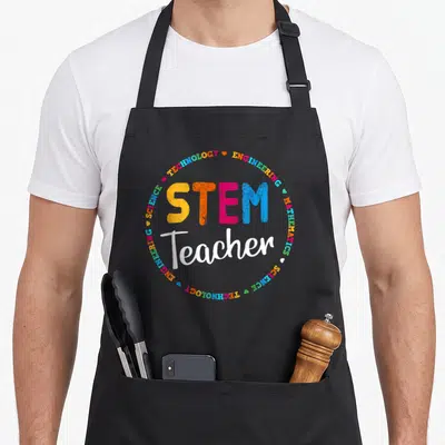 Outside teacher apron - STEM Teacher text surrounded by circular border of subject names and hearts in multicolor — shown on a black cotton canvas apron against a white background