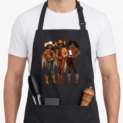 African western aprons - Three cartoon women in cowboy hats and fringe jackets standing in brown and tan — shown on a black cotton canvas apron against a white background