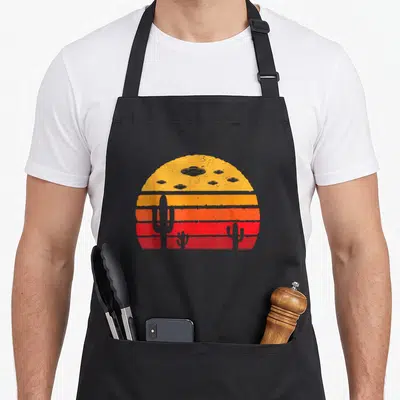Ufo weaving apron - Retro sunset with UFOs and three cacti in yellow orange red stripes — shown on a black cotton canvas apron against a white background