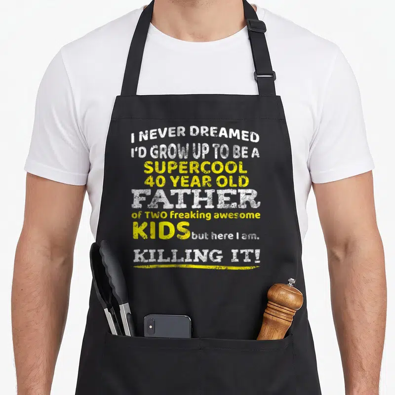 Father 40th birthday apron - White and yellow stacked typography reading I never dreamed I'd grow up to be a supercool — shown on a black cotton canvas apron against a white background