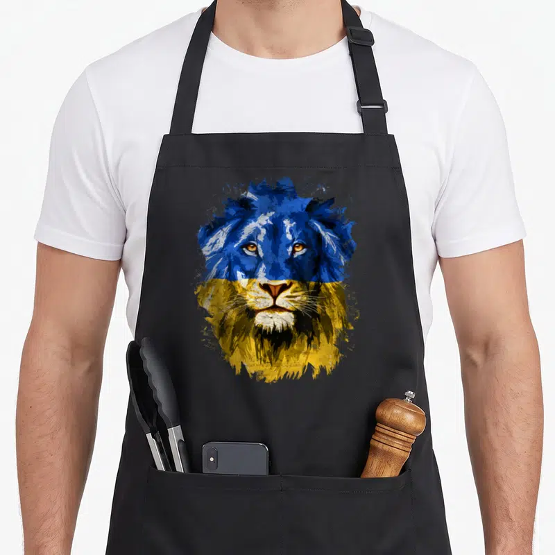Ukraine face apron - Realistic lion head in blue and yellow splatter art with black details — shown on a black cotton canvas apron against a white background