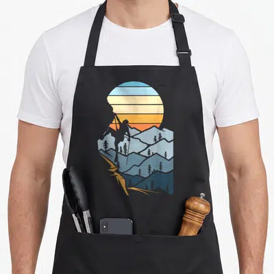 Mountain hobby apron - Retro hiker silhouette climbing blue mountains under a striped orange and yellow sun — shown on a black cotton canvas apron against a white background