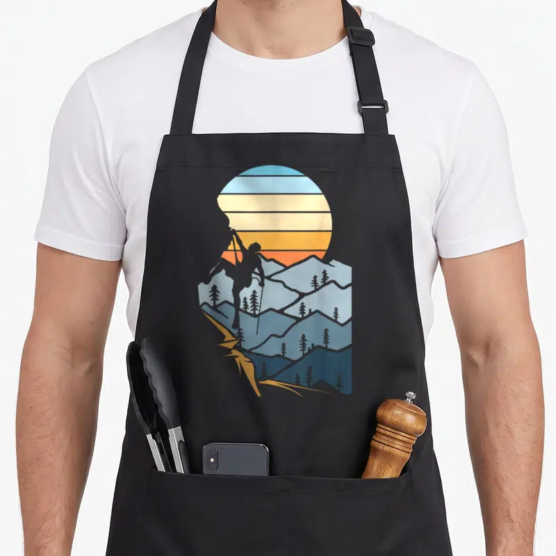 Mountain hobby apron - Retro hiker silhouette climbing blue mountains under a striped orange and yellow sun — shown on a black cotton canvas apron against a white background