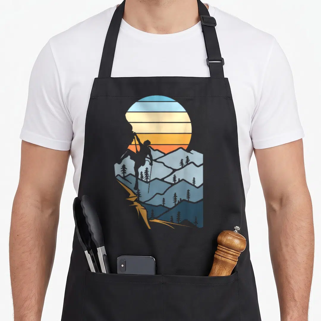 Mountain hobby apron - Retro hiker silhouette climbing blue mountains under a striped orange and yellow sun — shown on a black cotton canvas apron against a white background