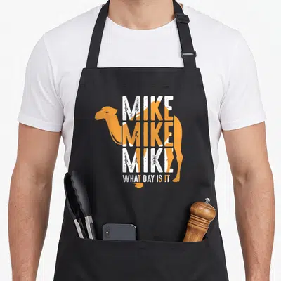Mike camel apron - Orange camel silhouette with stacked white distressed text reading MIKE MIKE MIKE WHAT DAY IS IT — shown on a black cotton canvas apron against a white background