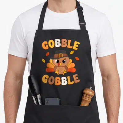 Toddler thanksgiving day aprons - Cartoon turkey in pilgrim hat with GOBBLE text and autumn leaves — shown on a black cotton canvas apron against a white background