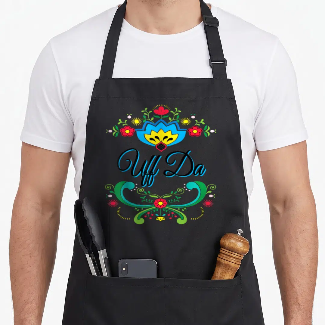 Uff norwegian kitchen aprons - Uff Da text between two symmetrical floral ornaments in red blue yellow green and black — shown on a black cotton canvas apron against a white background