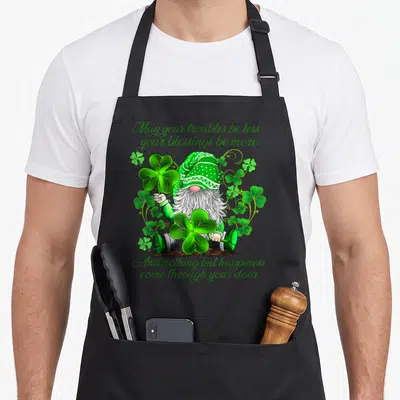 Christian st patrick day aprons - Cartoon gnome holding shamrocks with Irish blessing text in green script — shown on a black cotton canvas apron against a white background