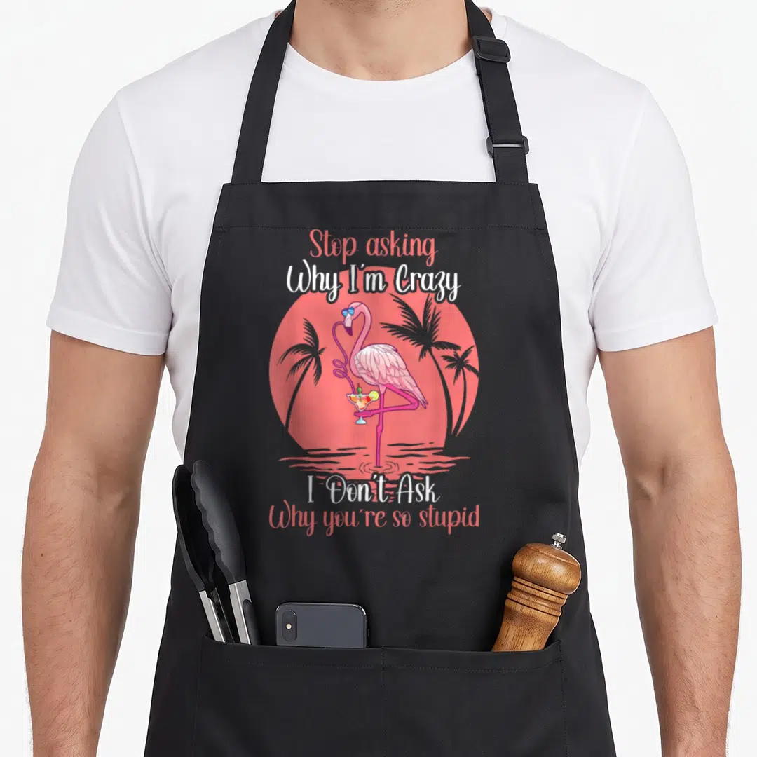 Stop asking flamingo apron - Pink flamingo with cocktail inside red sun surrounded by palm trees and split text — shown on a black cotton canvas apron against a white background