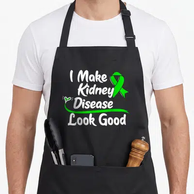 Survivor kidney disease awareness apron - I Make Kidney Disease Look Good text with green ribbon and heart swirl — shown on a black cotton canvas apron against a white background