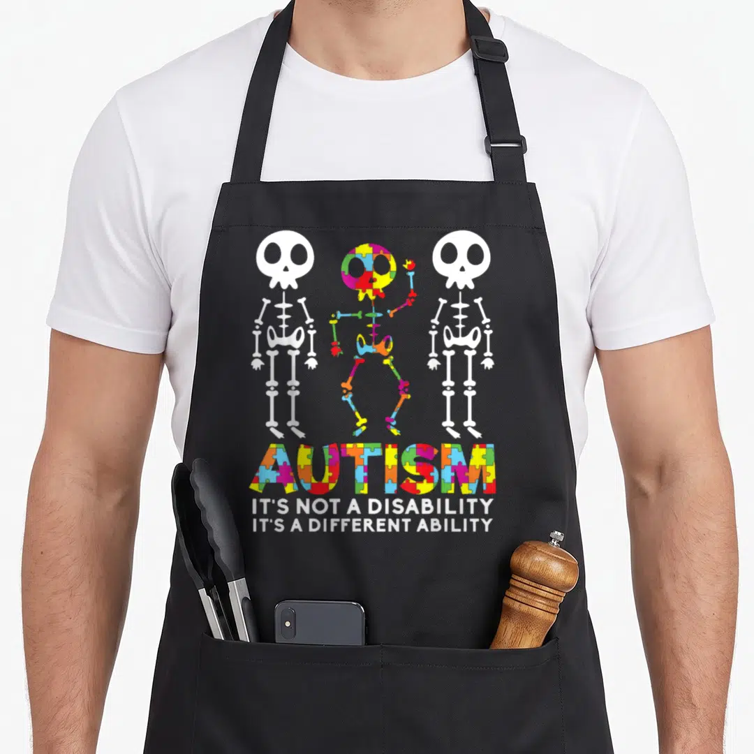 Dance skull apron - Three skeletons above colorful puzzle piece text reading autism its not a disability — shown on a black cotton canvas apron against a white background