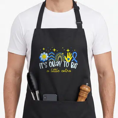 Syndrome an extra apron - IT'S OKAY TO BE a little extra text with blue yellow flower hand and ribbon symbols — shown on a black cotton canvas apron against a white background