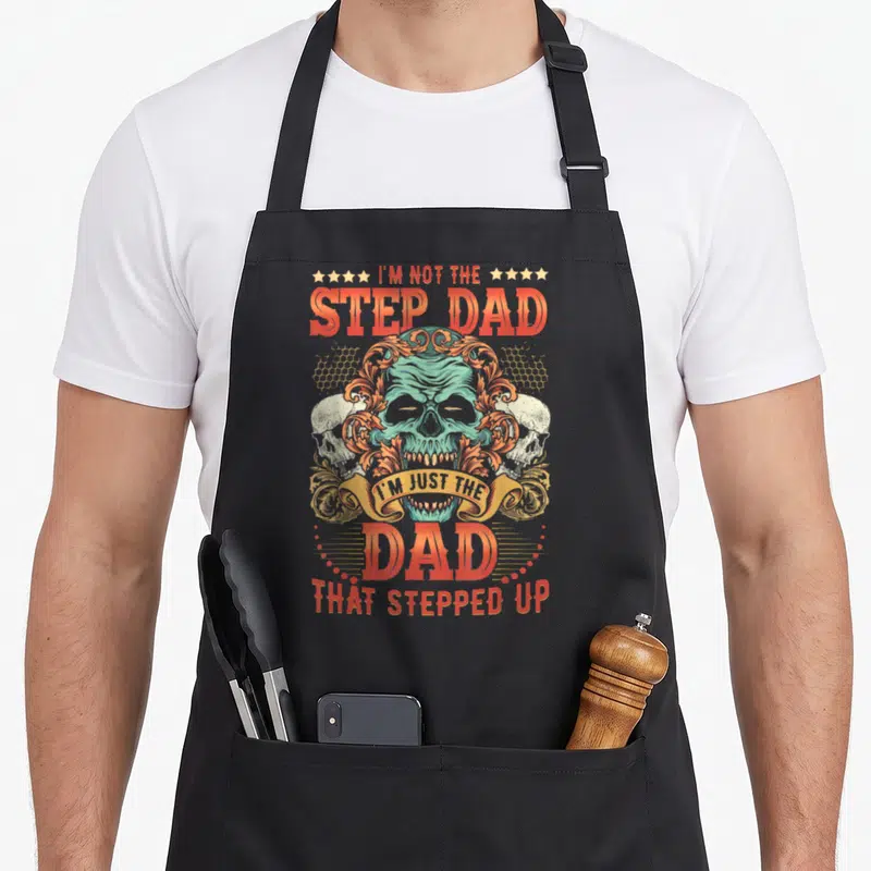 Funny stepped stepdad apron - Teal skull with orange swirls and two beige skulls under text about being a dad that — shown on a black cotton canvas apron against a white background