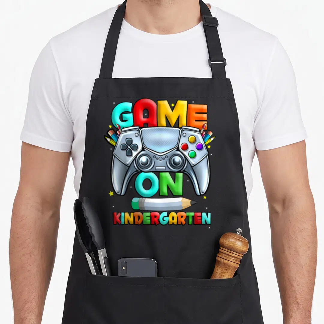 School game kindergarten apron - Silver game controller with pencils and paintbrushes surrounding multicolored GAME — shown on a black cotton canvas apron against a white background