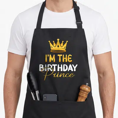 Retro prince birthday apron - Gold crown above stacked I'M THE BIRTHDAY Prince text in gold and white — shown on a black cotton canvas apron against a white background