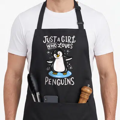 Retro penguin aprons - Cartoon penguin on blue ice with white text reading JUST A GIRL WHO LOVES PENGUINS — shown on a black cotton canvas apron against a white background