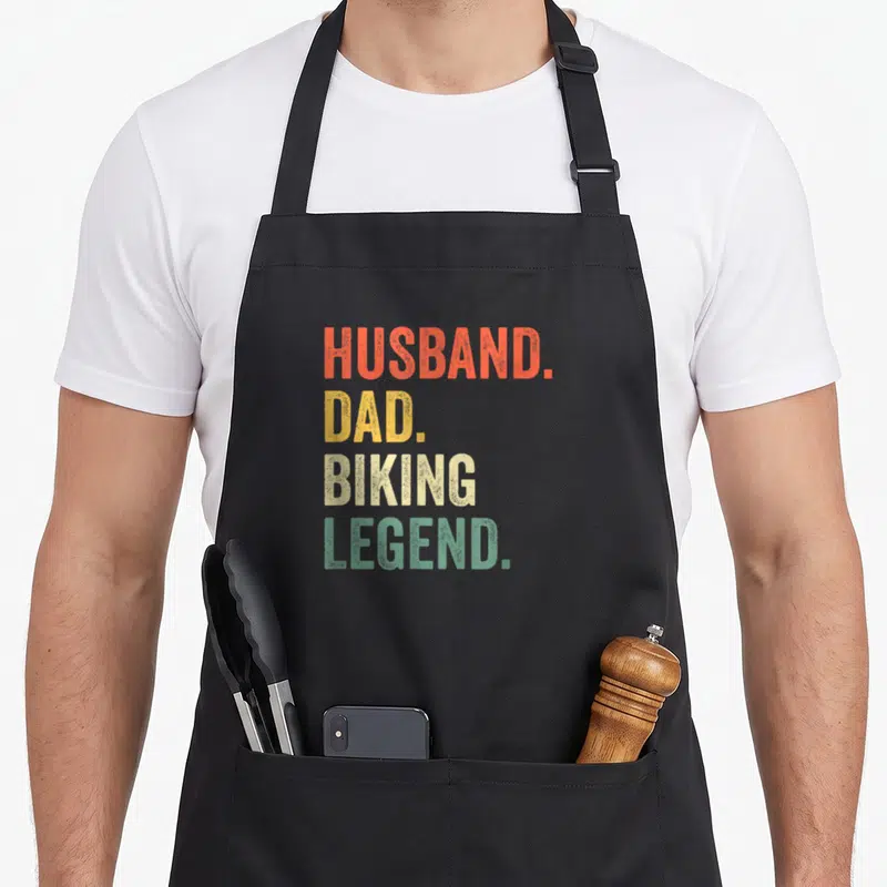 Bike husband apron - Stacked text HUSBAND. DAD. BIKING. LEGEND. in orange yellow white and green — shown on a black cotton canvas apron against a white background