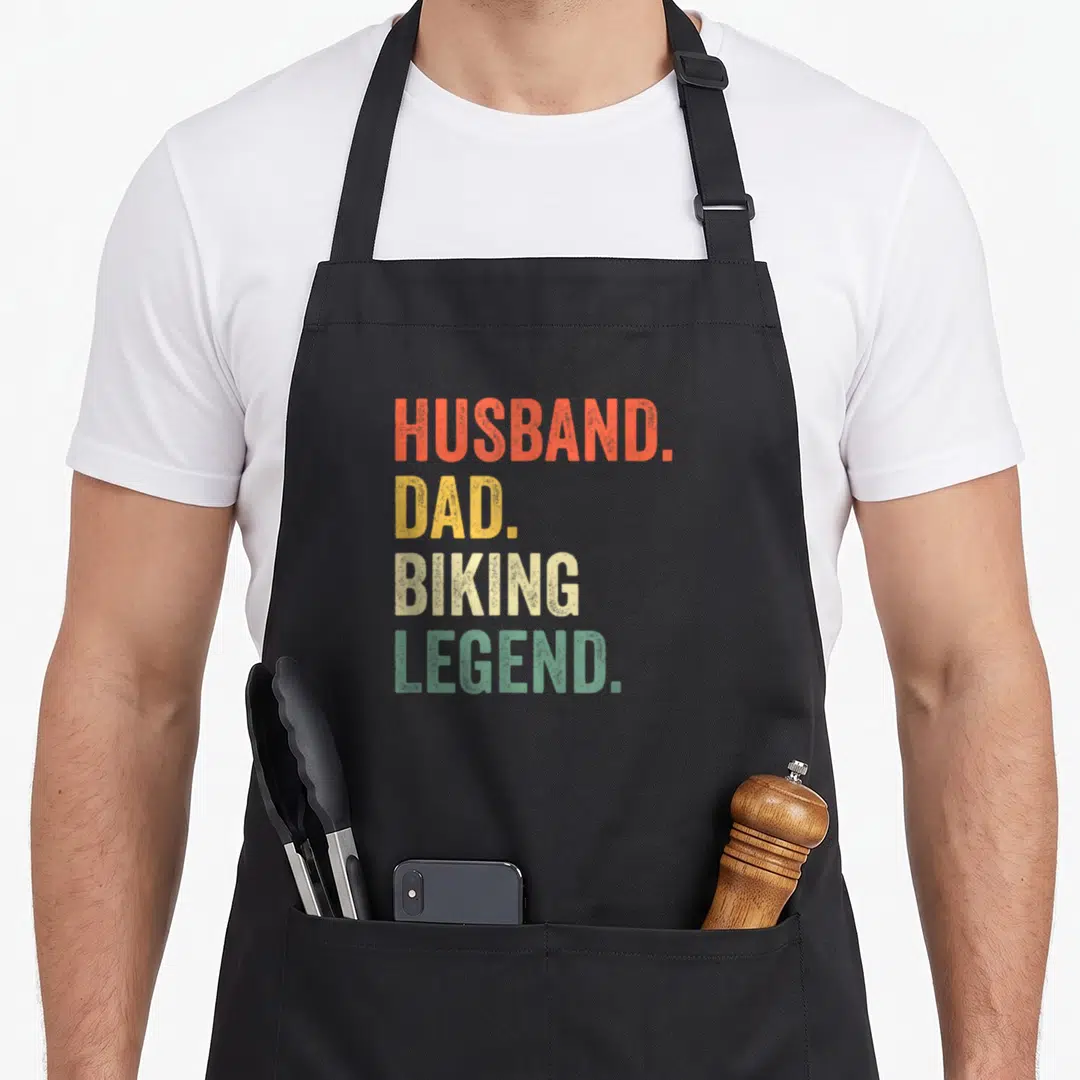 Bike husband apron - Stacked text HUSBAND. DAD. BIKING. LEGEND. in orange yellow white and green — shown on a black cotton canvas apron against a white background