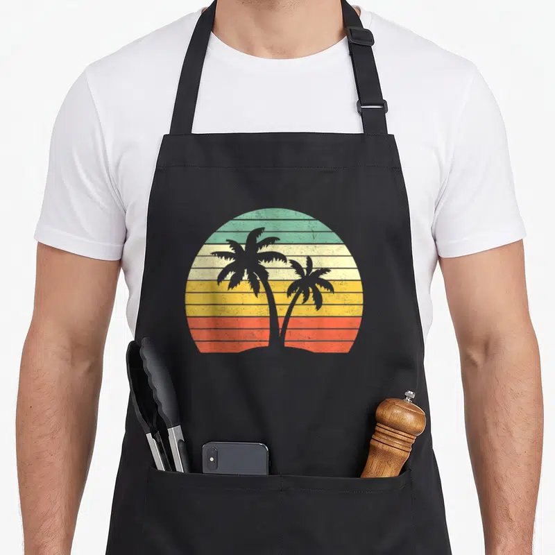 Tree tropical print aprons - Two grey palm trees in front of a teal yellow orange red striped sun — shown on a black cotton canvas apron against a white background