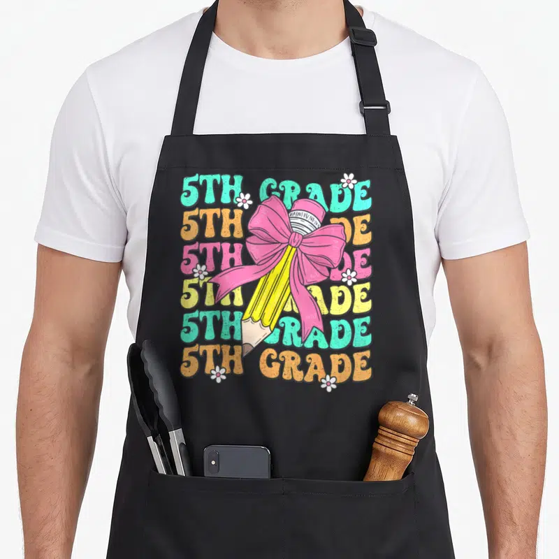 School groovy grade apron - Yellow pencil with pink bow over six rows of 5th Grade text in teal orange pink and yellow — shown on a black cotton canvas apron against a white background