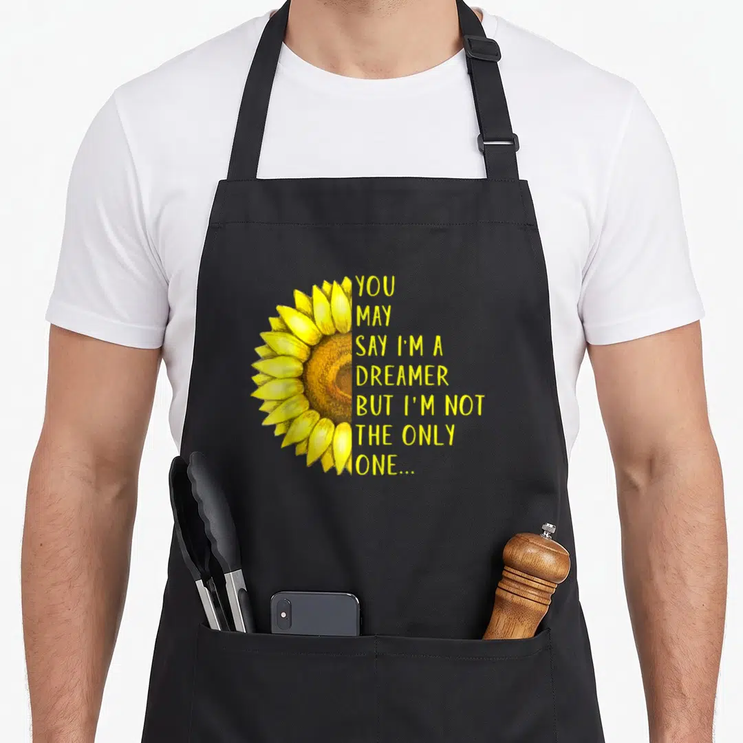 But dreamer apron - Half yellow watercolor sunflower beside stacked yellow text on black background — shown on a black cotton canvas apron against a white background