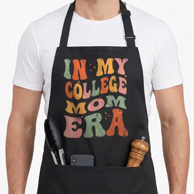 College mom era apron - Retro text IN MY COLLEGE MOM ERA with scattered stars in orange, yellow, green, and pink — shown on a black cotton canvas apron against a white background