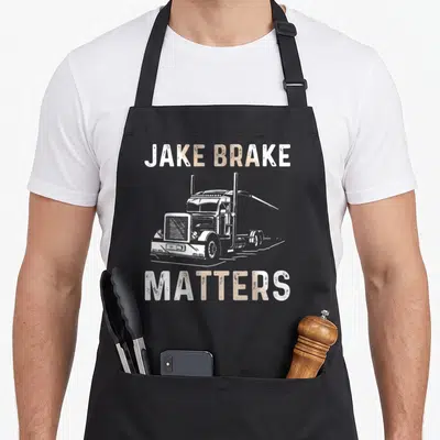 Driver trailer semi truck apron - White semi-truck illustration between JAKE BRAKE and MATTERS text in distressed tan — shown on a black cotton canvas apron against a white background