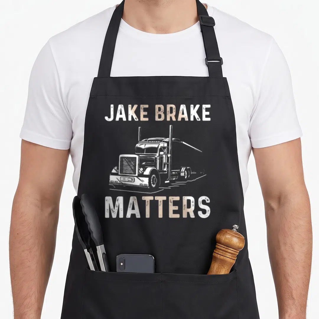 Driver trailer semi truck apron - White semi-truck illustration between JAKE BRAKE and MATTERS text in distressed tan — shown on a black cotton canvas apron against a white background