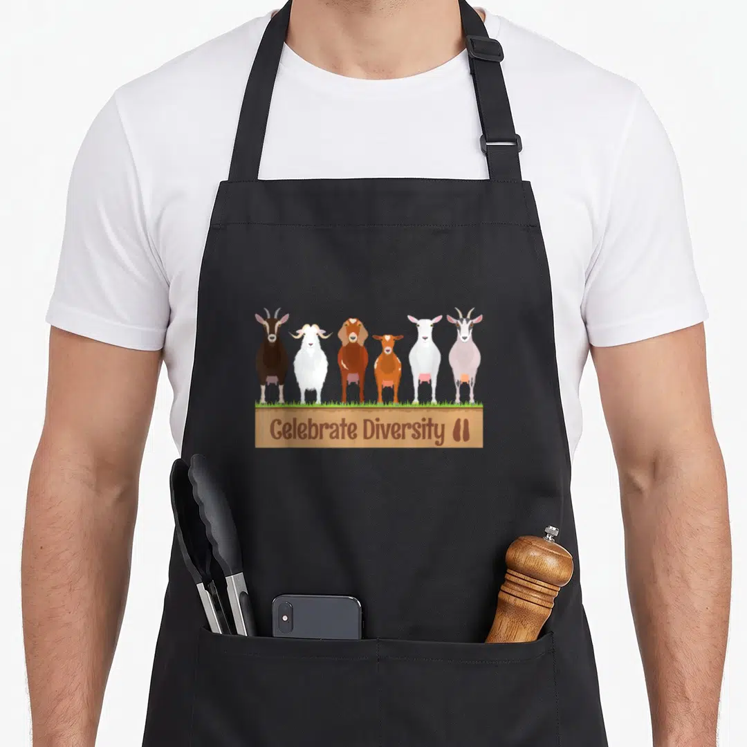 Celebrate farm apron - Six brown white and grey goats standing on grass above a wooden sign — shown on a black cotton canvas apron against a white background