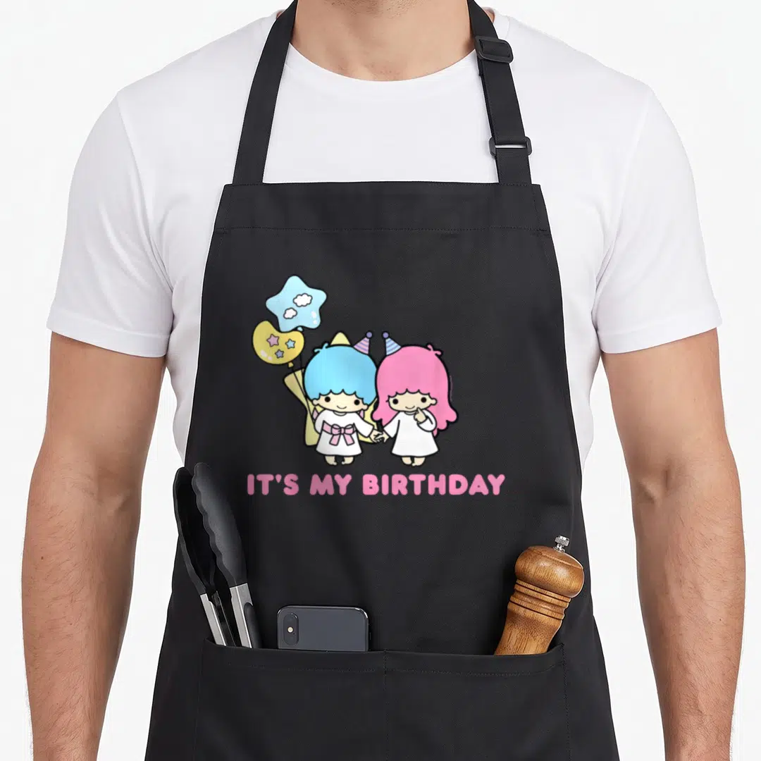 Star birthday apron - Two cartoon children holding hands with star and round balloons above pink IT'S MY BIRTHDAY text — shown on a black cotton canvas apron against a white background