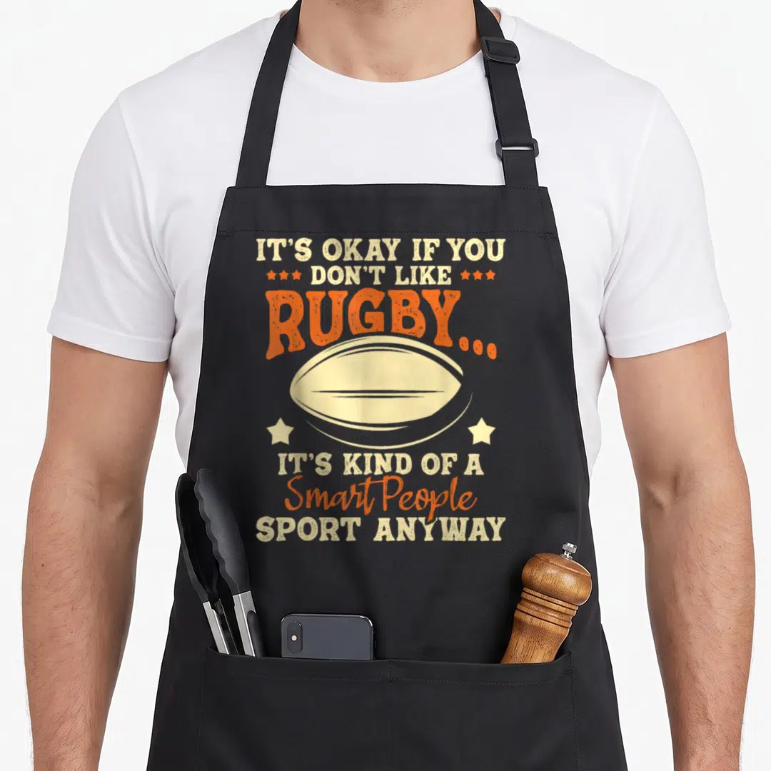 Okay rugby apron - Retro beige and orange rugby ball with text about it being a smart people sport surrounded by stars — shown on a black cotton canvas apron against a white background