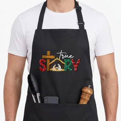 Story womens christmas apron - True Story text with wooden cross and nativity scene inside the letter O using red green — shown on a black cotton canvas apron against a white background
