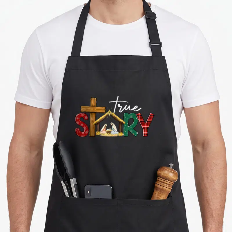 Story womens christmas apron - True Story text with wooden cross and nativity scene inside the letter O using red green — shown on a black cotton canvas apron against a white background