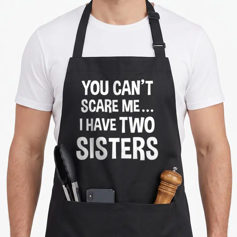 Scare nun apron - White text reading You Can't Scare Me I Have Two Sisters on black background — shown on a black cotton canvas apron against a white background