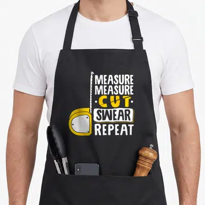 Measure woodworking apron - Yellow tape measure with extended white tape and stacked text MEASURE CUT SWEAR REPEAT — shown on a black cotton canvas apron against a white background