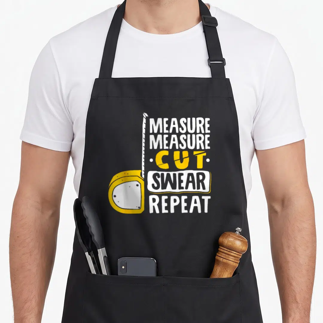 Measure woodworking apron - Yellow tape measure with extended white tape and stacked text MEASURE CUT SWEAR REPEAT — shown on a black cotton canvas apron against a white background