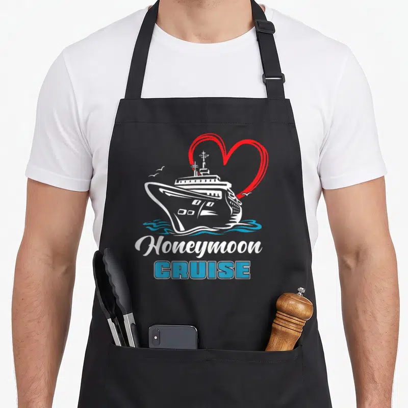 Honeymoon newlywed aprons - White cruise ship with red heart outline and blue Honeymoon CRUISE text — shown on a black cotton canvas apron against a white background
