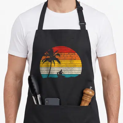 Surfer surfing player apron - Retro striped sunset with palm tree and surfer silhouette in red orange yellow teal grey — shown on a black cotton canvas apron against a white background