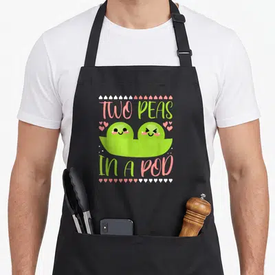 Peas kawaii apron - Cartoon two green peas in a pod with pink and white hearts and text — shown on a black cotton canvas apron against a white background
