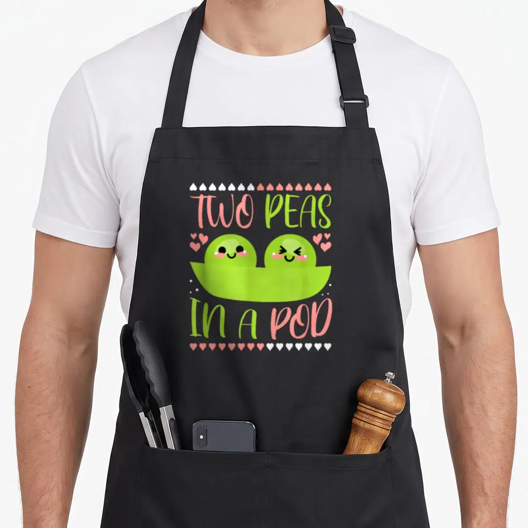 Peas kawaii apron - Cartoon two green peas in a pod with pink and white hearts and text — shown on a black cotton canvas apron against a white background