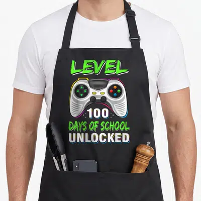 100th school video game apron - Cartoon game controller with LEVEL 100 DAYS OF SCHOOL UNLOCKED text in green and white — shown on a black cotton canvas apron against a white background