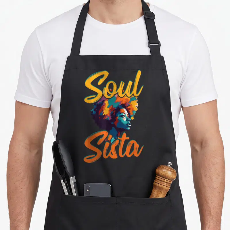 Cute funk 70s apron - Soul Sista text framing colorful cartoon woman profile with large afro in yellow orange blue — shown on a black cotton canvas apron against a white background