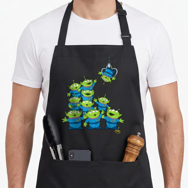 Claw alien apron - Green aliens in blue suits piled up with silver claw lifting one — shown on a black cotton canvas apron against a white background