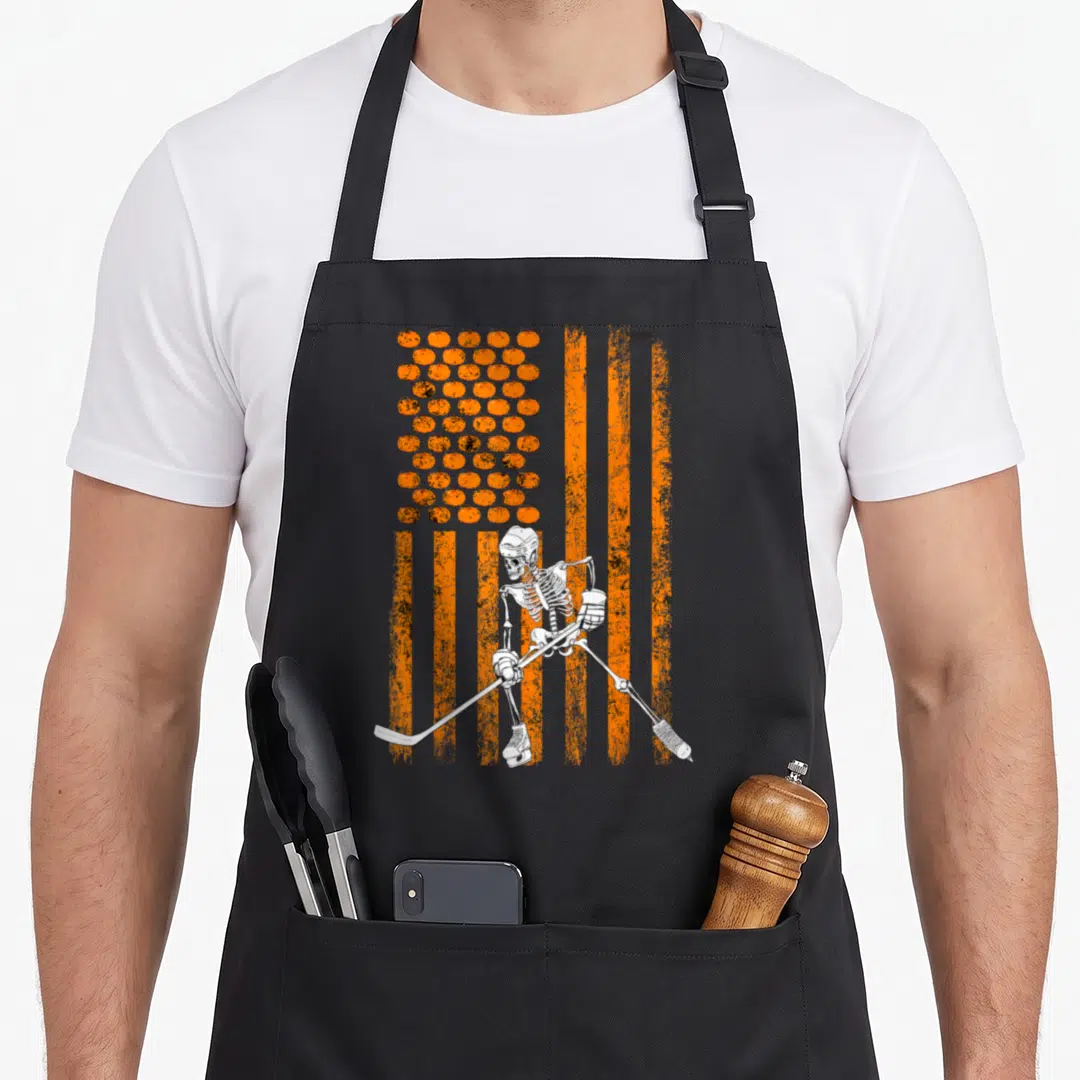 Player halloween apron - Skeleton hockey player in front of orange pumpkin and stripe flag background — shown on a black cotton canvas apron against a white background