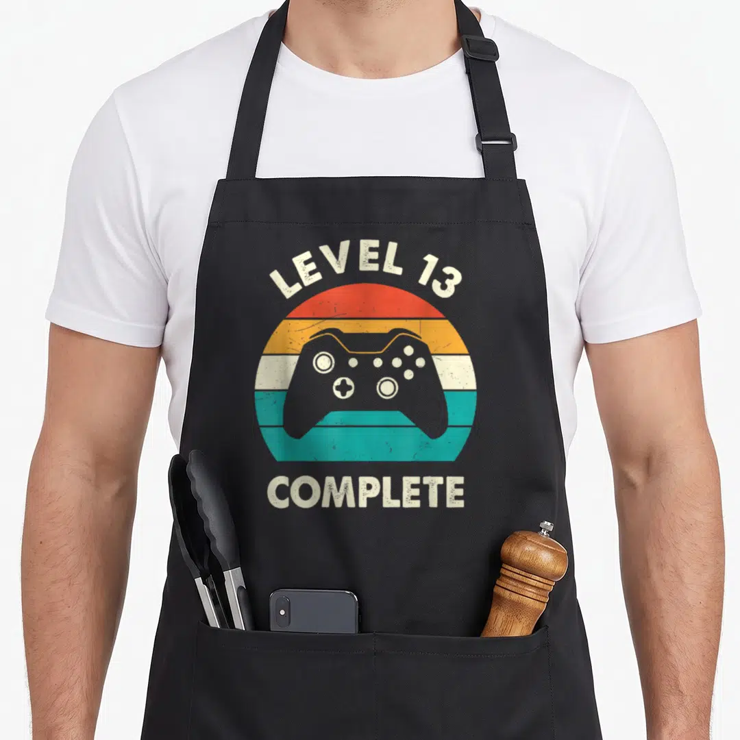 Level wedding aprons - Retro striped circle with game controller and LEVEL 13 COMPLETE text — shown on a black cotton canvas apron against a white background