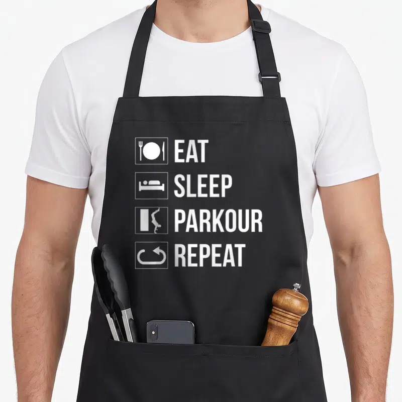 Sleep parkour apron - White icons showing a plate, bed, wall climb, and refresh symbol next to matching text — shown on a black cotton canvas apron against a white background